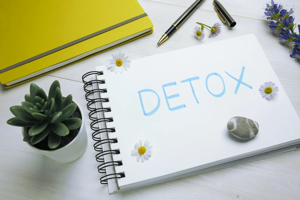Spiral notebook with the word detox surrounded by flowers, pens, and a small plant